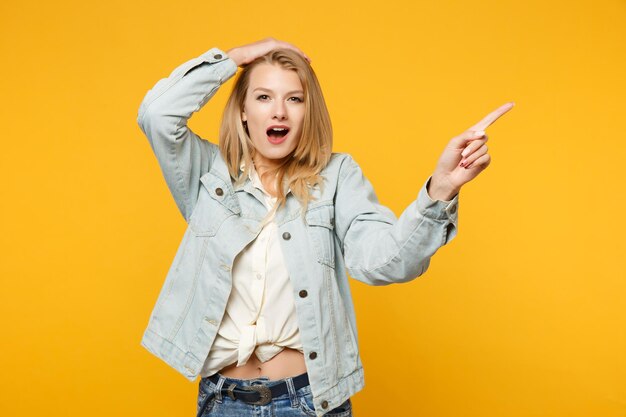 Portrait of shocked young woman in denim casual clothes pointing index finger aside, putting hand on head isolated on yellow orange background in studio. people lifestyle concept. mock up copy space