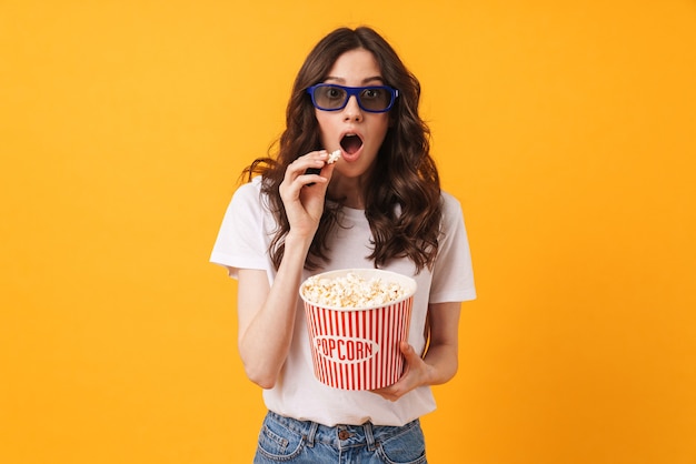 Il ritratto della giovane donna colpita che posa isolata sopra la parete gialla mangia il film dell'orologio del popcorn.