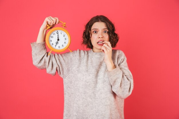 Portrait of a shocked young woman standing isolated over pink, showing alarm clock