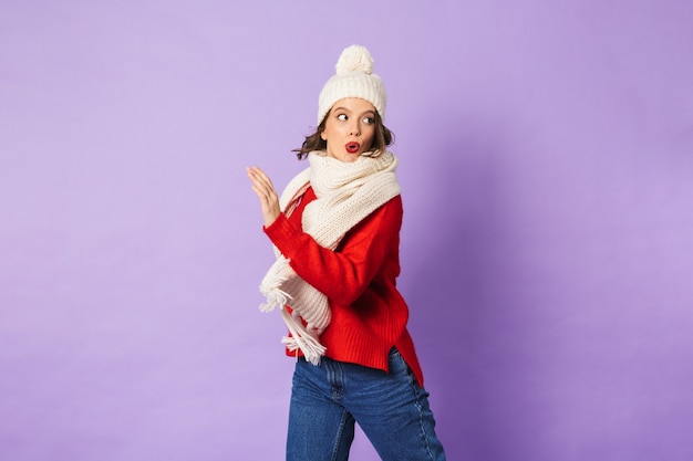 Portrait of a shocked young woman wearing winter hat isolated over purple wall.