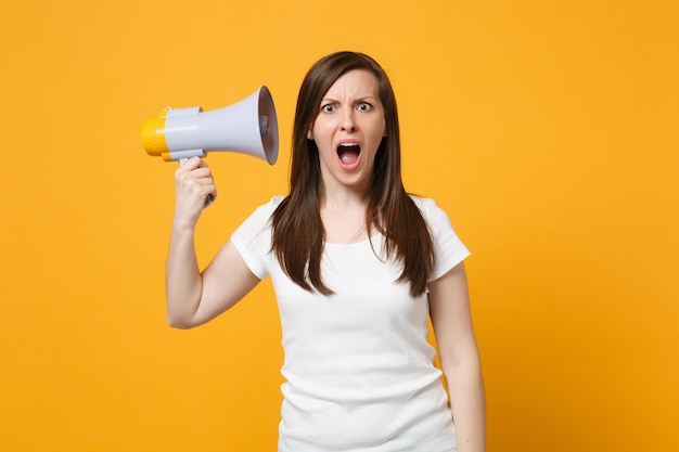 Portrait of shocked young woman in white casual clothes keeping mouth open, hold megaphone near ear isolated on bright yellow orange background in studio. People lifestyle concept. Mock up copy space.