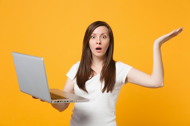 Portrait of shocked young woman in white casual clothes spreading hands, using laptop pc computer isolated on bright yellow orange background in studio. People lifestyle concept. Mock up copy space.