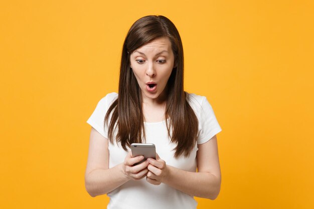 Portrait of shocked young woman in white casual clothes using mobile phone, typing sms message isolated on bright yellow orange wall background in studio. People lifestyle concept. Mock up copy space.