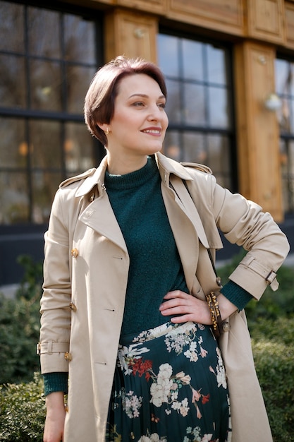 Portrait shooting of a attractive stylish short cut woman, Girl walks in the city outdoors. Stylish modern and feminine image, style. Girl in a beige cloak or coat and a green dress