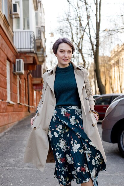 Portrait shooting of a attractive stylish short cut woman, Girl walks in the city outdoors. Stylish modern and feminine image, style. Girl in a beige cloak or coat and a green dress