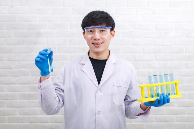 흰색 실험실 코트 고무 장갑과 안전 고글을 쓴 아시아 전문 남성 과학자가 벽돌 벽 배경에 손에 샘플 테스트 튜브 랙을 들고 있는 카메라를 바라보며 웃고 있습니다.