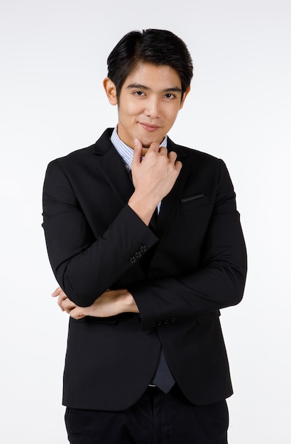 Portrait shot of Asian young handsome happy intelligence successful businessman in black formal suit stand hold hand touch chin brooding thinking and smiling alone in front of white wall background.