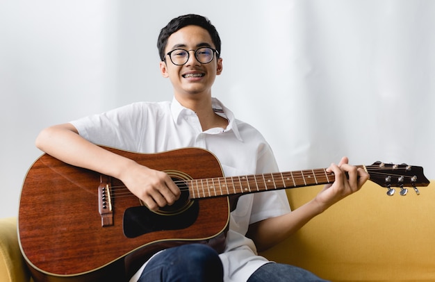 Portrait shot of attractive smiling young male teenager\
enjoying playing the acoustic guitar. junior guitarist in white\
clothes sitting on couch and holding an instrument while looking at\
the camera