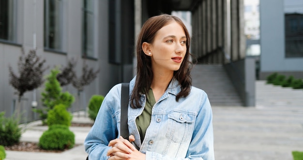 Portrait shot of Caucasian pretty young pretty woman in blue jeans jacket with bag standing at city street and smiling joyfully to camera. Beautiful happy cheerful female outdoors.