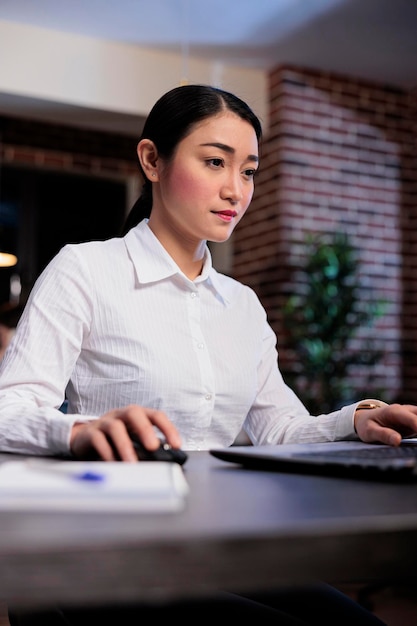 Portrait shot of confident businesswoman at night sitting in office workspace while working on laptop. Management department leader analyzing financial charts and graphs while checking documentation.