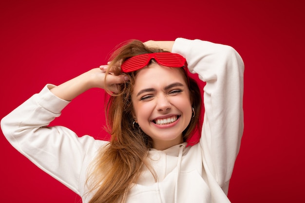 Portrait shot of fascinating positive young blonde woman isolated over colourful background wearing casual white hoodie and stylish sunglasses.