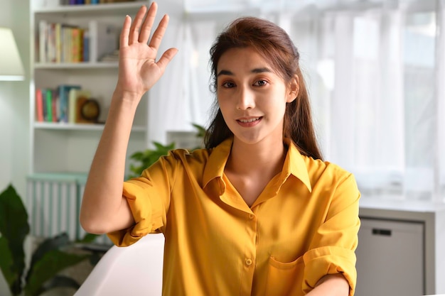 Portrait shot of happy friendly and confident Asian businesswoman sitting on a couch at home, looking and talking at camera and waving hand saying hello during video call