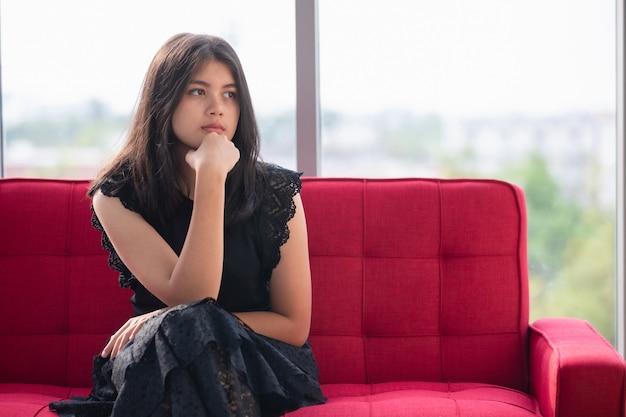 Portrait shot of junior young Thai-Turkish teenager wearing trendy dress sitting on the red couch and posing hand to chin gesture in the studio. Thoughtful girl thinking about the college project