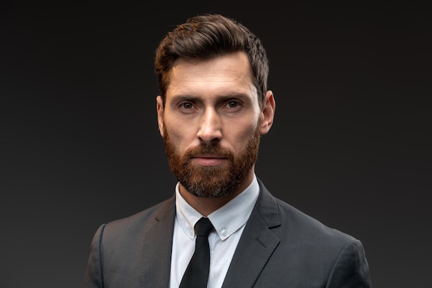 Portrait shot of middle aged bearded businessman wearing suit and tie looking at the camera with serious face while standing at isolated black background