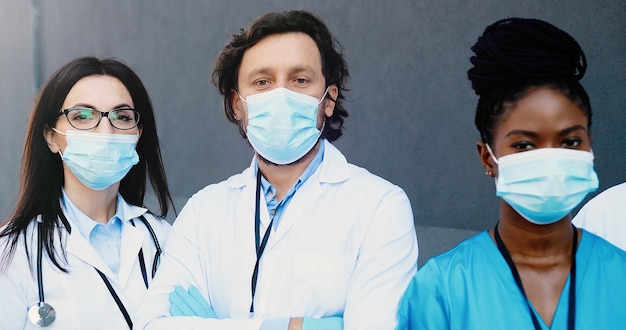 Portrait shot of mixed-races young males and females doctors in medical masks and white gowns standing and looking at camera. Multi-ethnic medics, men and women. Coronavirus concept.