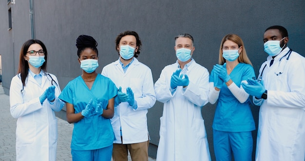 Foto ritratto di giovani medici uomini e donne di razza mista sparati in maschere mediche, guanti e abiti bianchi in piedi, applaudendo e guardando la fotocamera. medici multietnici. concetto di vittoria covid-19. applausi.