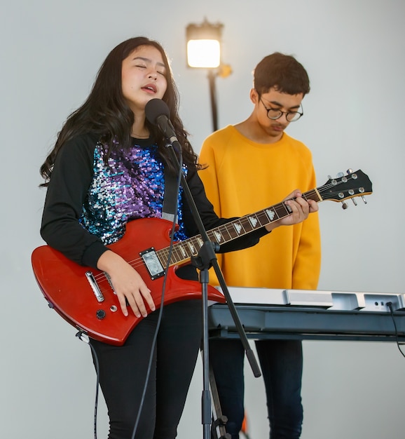 Portrait shot of a young teenage singer singing a song with a microphone and playing the electric guitar. Professional junior student vocalist practicing with a male pianist in the background.