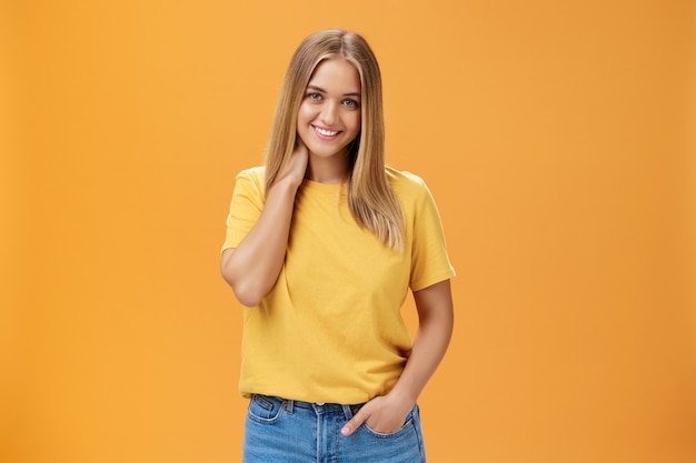 Portrait of shy and timid girl with tan and straight fait hair rubbing neck and smiling holding hand in pocket posing against orange background