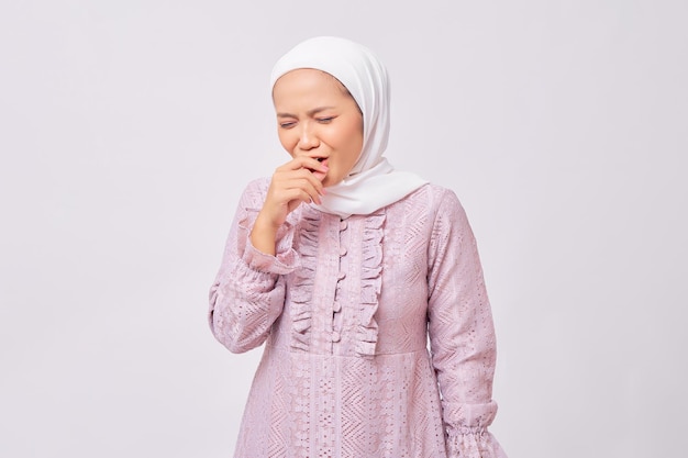 Portrait of sick beautiful young Asian Muslim woman wearing hijab and purple dress feeling unwell and coughing isolated on white studio background