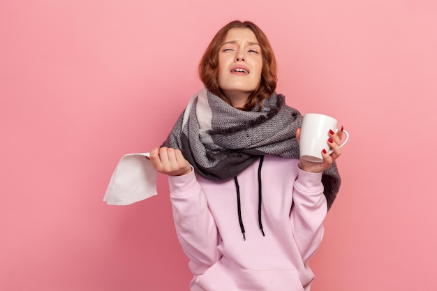 Portrait of sick curly haired teenage girl in hoodie wrapped in scarf crying holding cup of tea and napkin treatment Indoor studio shot isolated on pink background