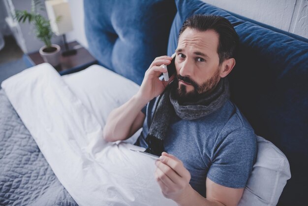 Photo portrait of sick man holding thermometer calling a doctor and looking at the camera while sitting on the bed