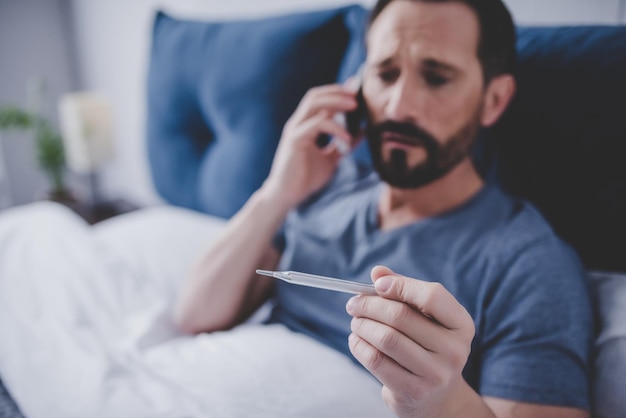 Portrait of sick man holding thermometer and calling a doctor while sitting on the bed