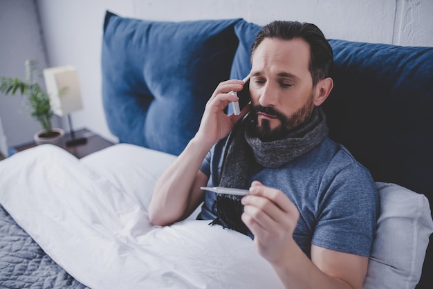 portrait of sick man holding thermometer and calling a doctor while sitting on the bed