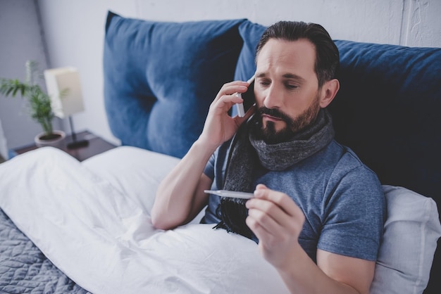 Photo portrait of sick man holding thermometer and calling a doctor while sitting on the bed