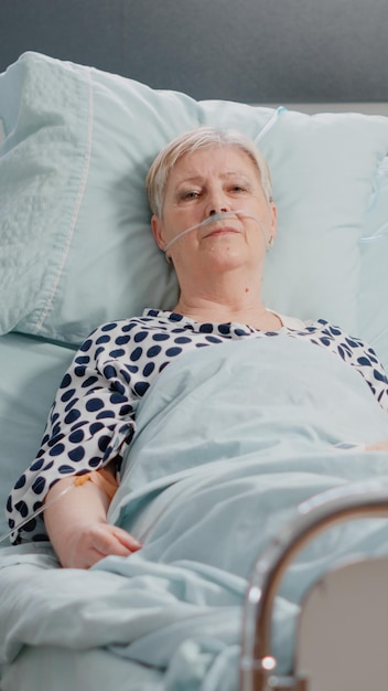 Portrait of sick patient laying in hospital ward bed, looking\
at camera. senior woman with nasal oxygen tube and iv drip bag\
resting to cure diagnosis for healthcare and recovery.