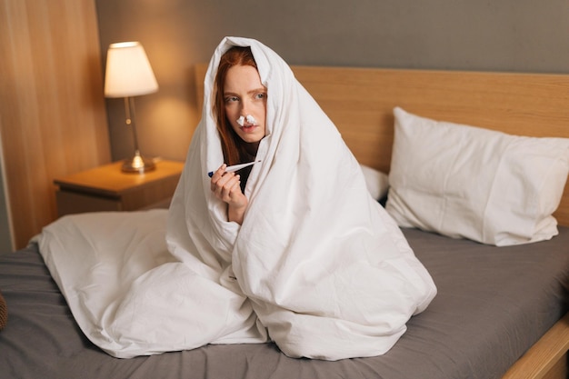 Portrait of sick sad woman suffering from runny nose disease paper tissues inserted into nose measuring temperature with thermometer sitting in bed