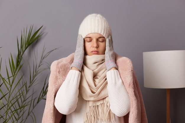 Portrait of sick unhealthy frozen woman wearing cap and coat posing in living room feels pain in head catching cold in cold season massaging her temples