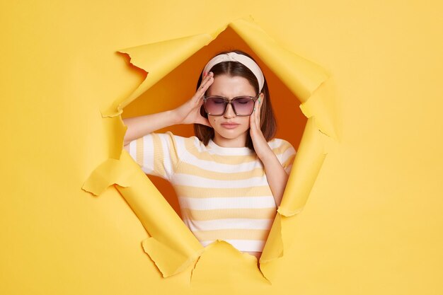 Photo portrait of sick unhealthy woman wearing striped tshirt hair band and sunglasses breaking through paper hole in yellow wall suffering headache being ill