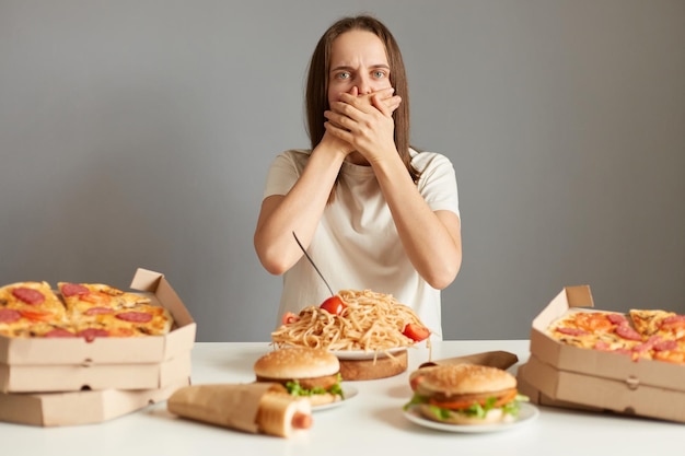 Портрет больной женщины с каштановыми волосами в белой футболке, сидящей за столом на сером фоне и чувствующей тошноту после переедания нездоровой пищи