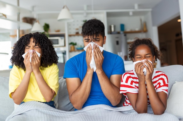 Portrait of sick young black family of three people blowing runny noses while sitting together on the sofa with napkins and covered with blanket