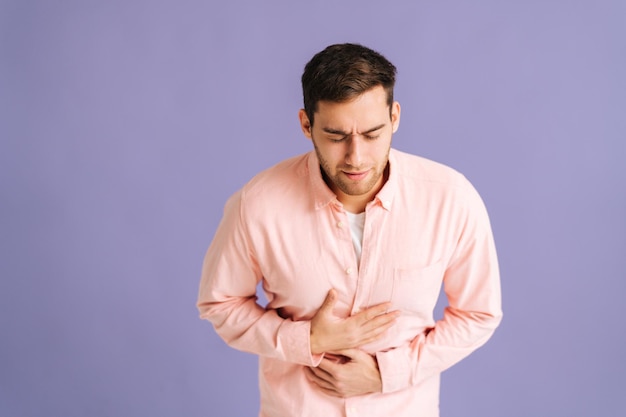 Portrait of sick young man feeling sudden stomach pain, severe abdominal distress, suffering nausea going to throw up in studio. Ill male put hands on abdomen suffering from stomach-ache.