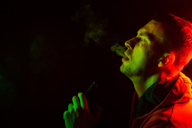 Portrait of a side mouth part of guys face with a colored backlight of green and red smoking a vape and exhaling smoke in different directions on a black isolated background Puffs harmful to health