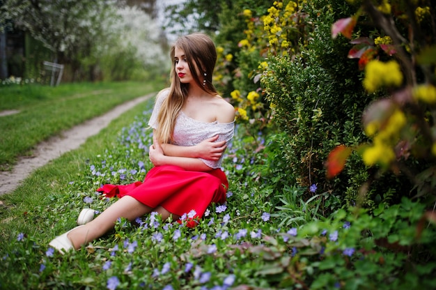 Portrait of sitiing beautiful girl with red lips at spring blossom garden on grass with flowers, wear on red dress and white blouse.