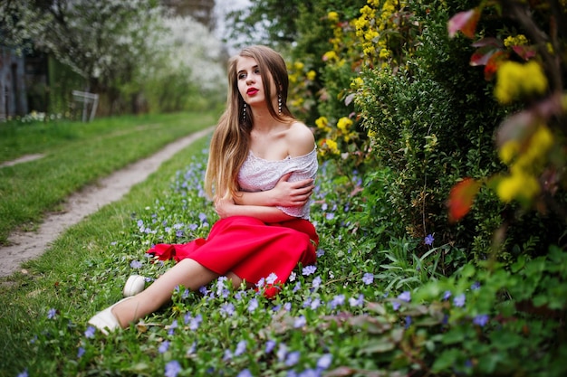 Portrait of sitiing beautiful girl with red lips at spring blossom garden on grass with flowers wear on red dress and white blouse