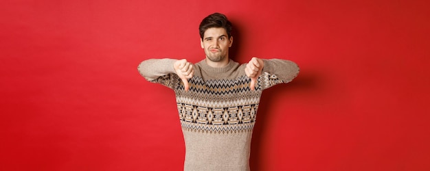 Portrait of skeptical and disappointed handsome man in christmas sweater dislike new year party showing thumbsdown express disapproval standing over red background