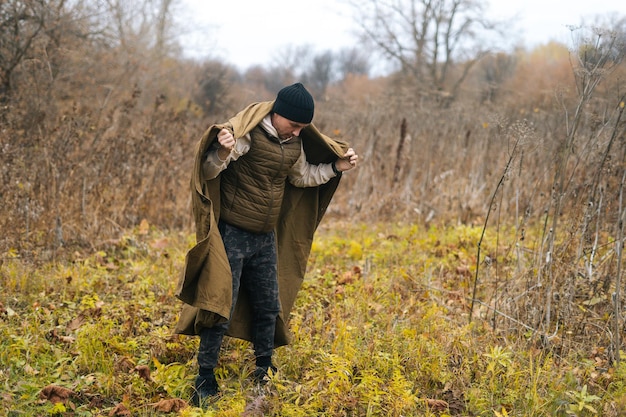 Portrait of skilled hiker man wearing green raincoat tent at outdoors on cloudy rainy day standing in thicket of bushes