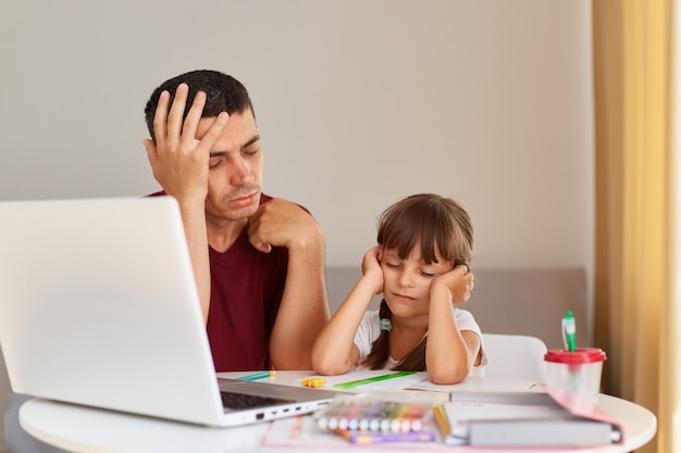 Photo portrait of sleeping daughter and father sitting at the desk while doing homework together, being tired of doing lots tasks, wearing casual style clothing, distant education.