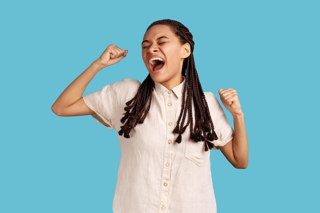 Portrait of sleepless woman with black dreadlocks yawning and raising hands up, feeling fatigued, standing with close eyes, wearing white shirt. Indoor studio shot isolated on blue background.