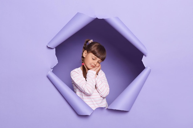 Portrait of sleepy tired adorable little girl with braids looking through breakthrough of purple background standing with closed eyes leaning on her palms feels sleepless