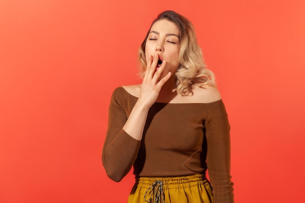 Portrait of sleepy tired blonde woman in brown blouse yawning and covering mouth with arm, feeling drowsy after sleepless night, lack of energy. Indoor studio shot isolated on red background
