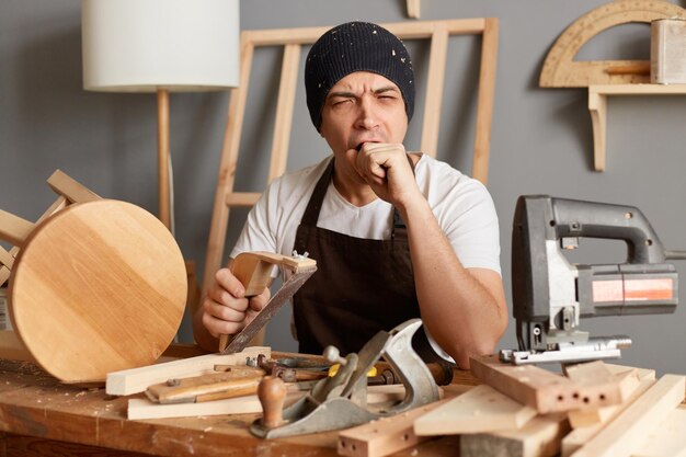 Portrait of sleepy tired exhausted young joiner wearing apron and cap working in workshop long hours yawning looks sleepy holding grinder in hands keeps eyes closed