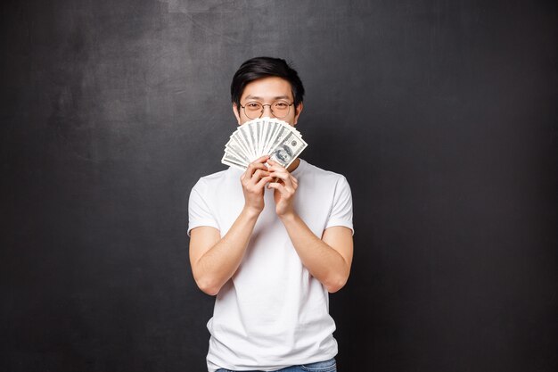 Portrait of sly happy and pleased asian young man winning big cash prize money, hiding face behind fan of dollars smiling with eyes, decide what to buy on it, standing 