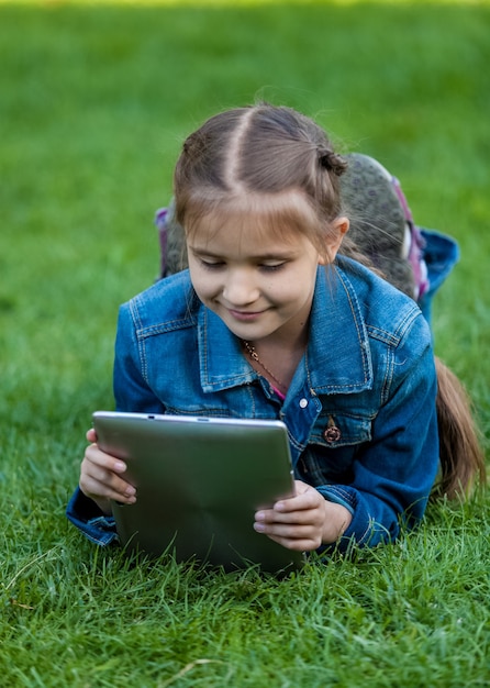 Портрет маленькой девочки, лежащей на траве и читающей книгу с планшета