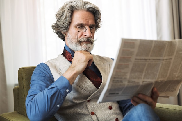 Portrait of smart adult businessman wearing stylish suit reading newspaper while sitting on armchair in hotel apartment