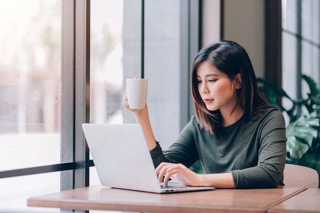 Portrait of smart asian woman freelance holding coffee cup and
working online with laptop in co-working space