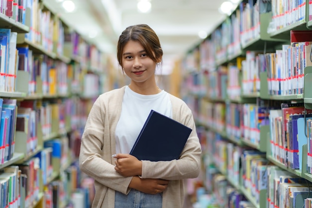 Портрет умного азиатского студента университета женщины, читающего книгу и смотрящего на камеру между книжными полками в библиотеке кампуса с copyspace.